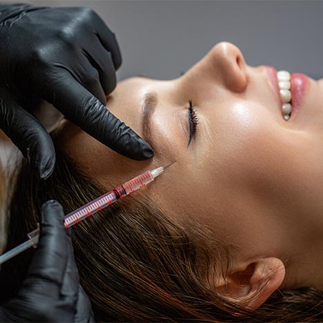 Gros plan d’une femme allongée sur le côté, les yeux fermés et légèrement souriante. Une personne portant des gants noirs tient une seringue près de la tempe et touche la peau du bout du doigt