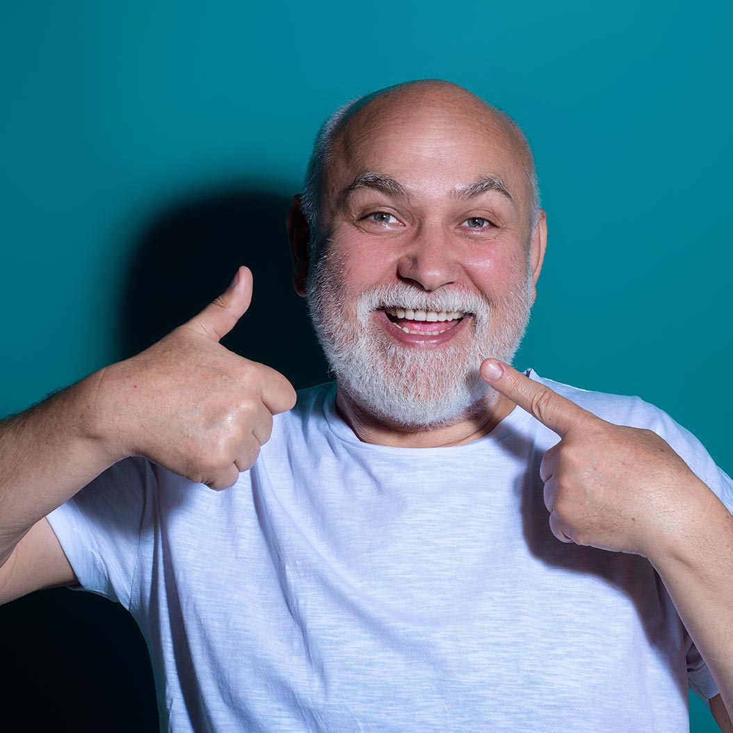 Portrait d’un homme souriant avec barbe blanche, pointant vers ses dents avec un pouce levé, sur fond bleu uni