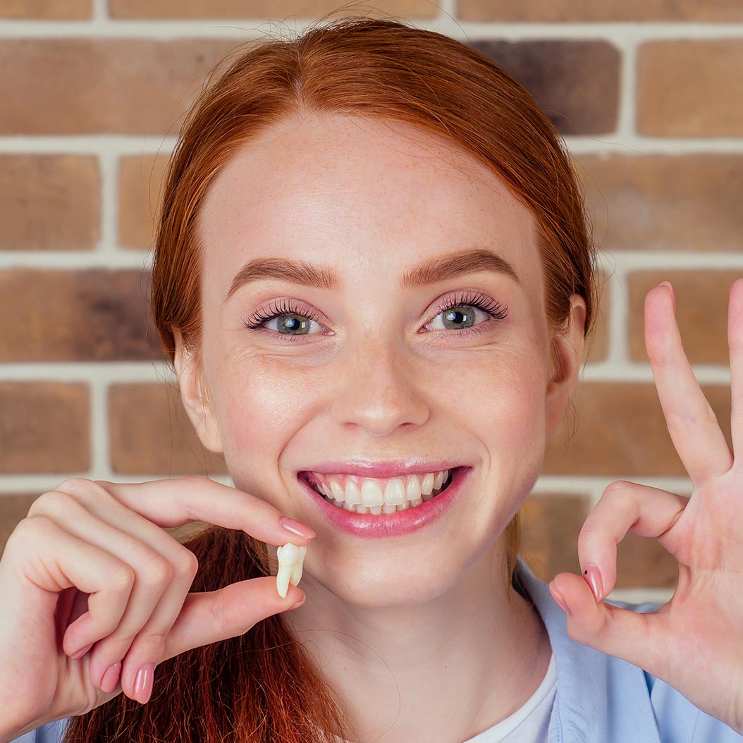 Jeune femme souriante devant un mur de briques, tenant une petite dent entre ses doigts et faisant un geste « OK » avec l’autre main.
