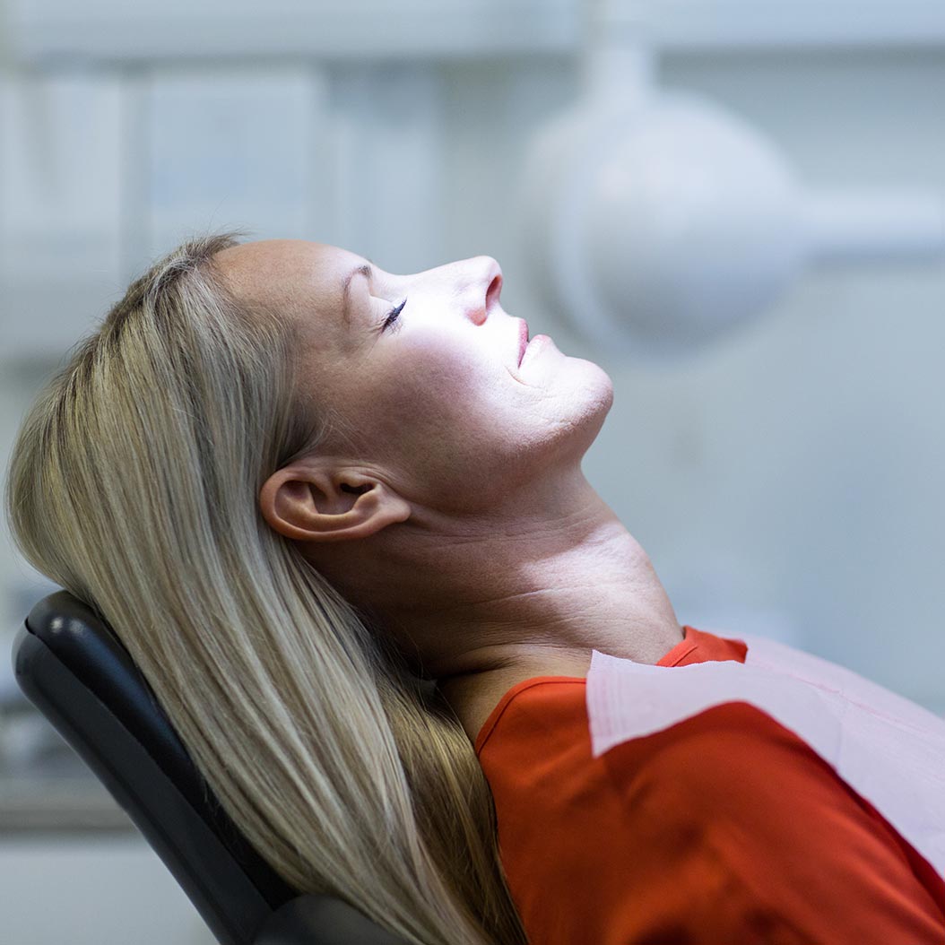 Femme assise dans un fauteuil dentaire, la tête inclinée vers l’arrière et les yeux fermés, éclairée par une lampe d’examen dans un cabinet clinique.